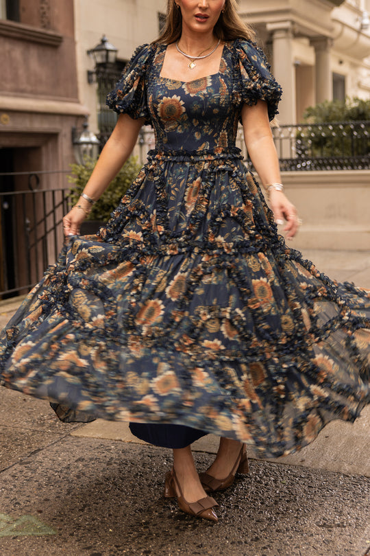 A woman in the Ivy City Co Marseille Dress in Sunflower Twilight, featuring blue floral patterns and ruffles, twirls on a wet city sidewalk with her face turned away. She pairs the dress with brown heels and gold jewelry, while classic residential buildings line the background.