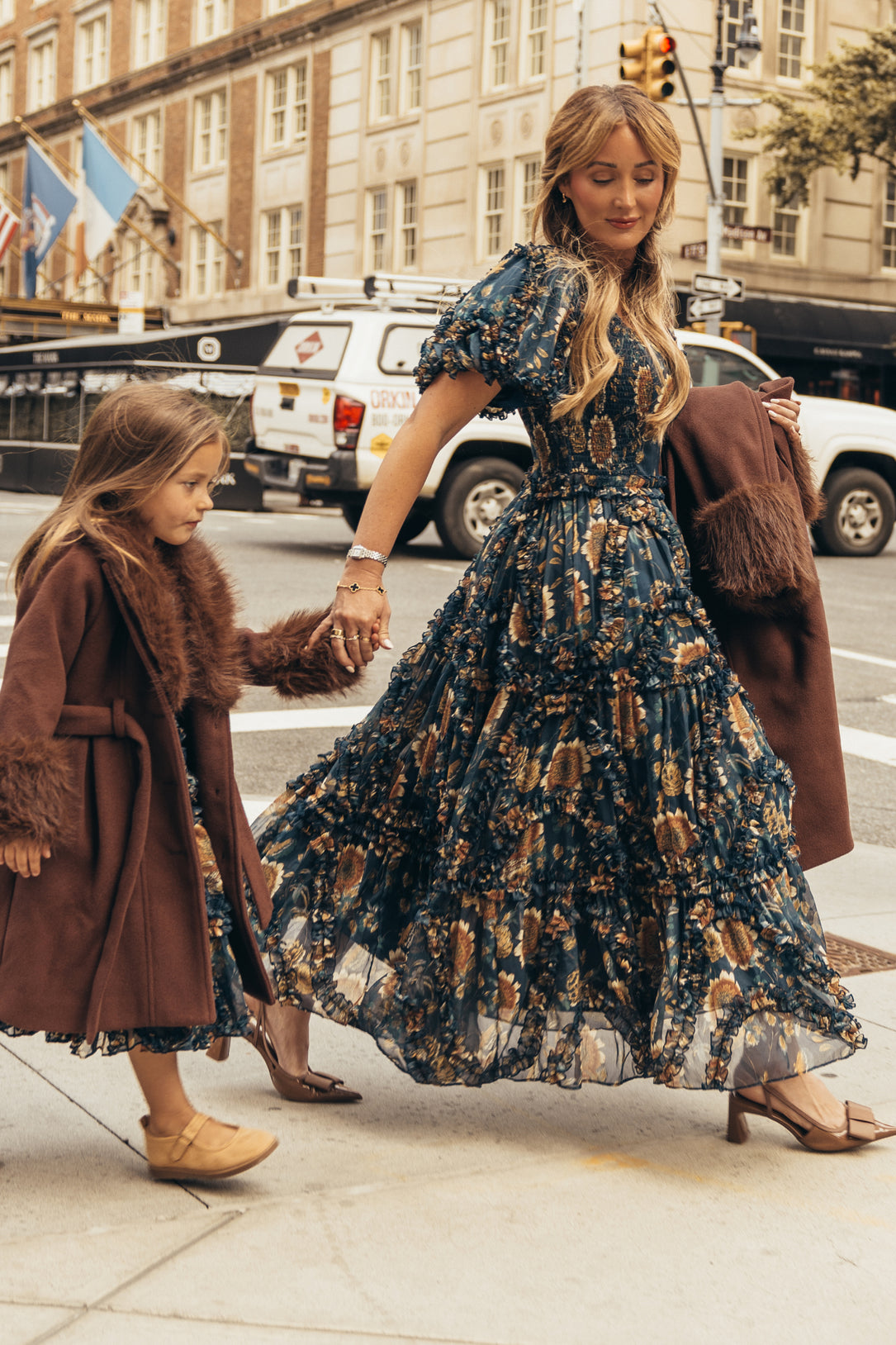 A woman wearing the Ivy City Co Marseille Dress in Sunflower Twilight—a flowy, ankle-length dress adorned with a beautiful floral pattern—walks hand-in-hand with a girl dressed in a brown coat with faux fur trim on a city sidewalk. The woman’s carried brown coat coordinates perfectly with the girl’s outerwear, creating a stylish matching look amidst the backdrop of buildings and cars.