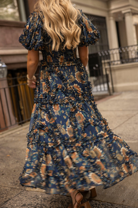A woman with long blonde hair walks on a city sidewalk, her back turned to the camera, wearing the Ivy City Co Marseille Dress in Sunflower Twilight—a flowing, floral-patterned navy dress with a square neckline.