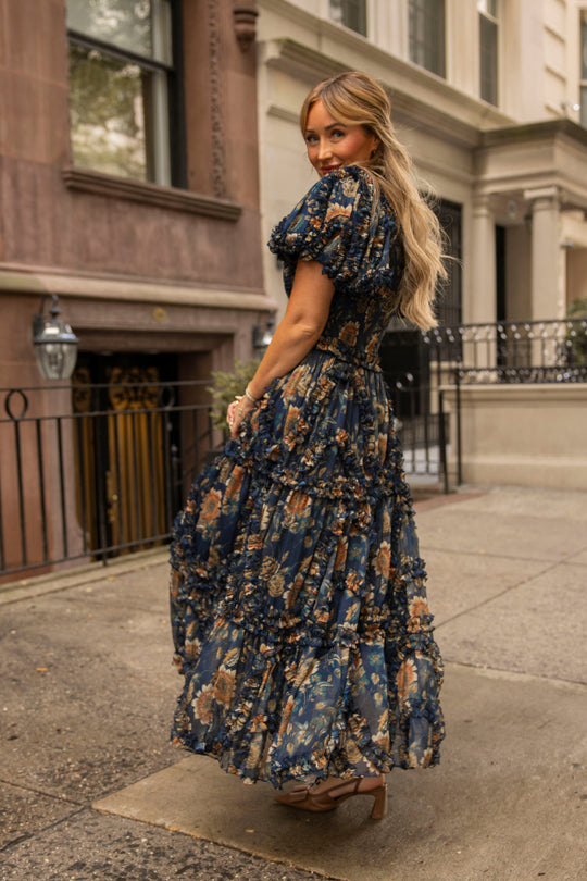 A woman with long blonde hair, wearing the Ivy City Co Marseille Dress in Sunflower Twilight—a navy blue floral dress with a square neckline—and heels stands on a city sidewalk, looking over her shoulder and smiling, with brownstone buildings in the background.
