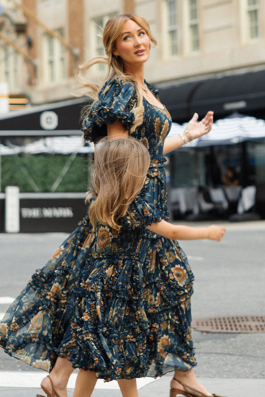 A woman and a young girl, both wearing the Ivy City Co Marseille Dress in Sunflower Twilight, walk hand in hand across a city street. The ankle-length flowy dresses move gracefully as they walk, with city buildings and outdoor seating visible in the background.