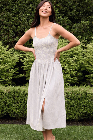 A woman stands outdoors before green bushes, smiling with her hands in the pockets of the Ivy City Co Maxwell Dress in Blue & White Pinstripe—a sleeveless smocked midi dress with thin straps and a front slit.