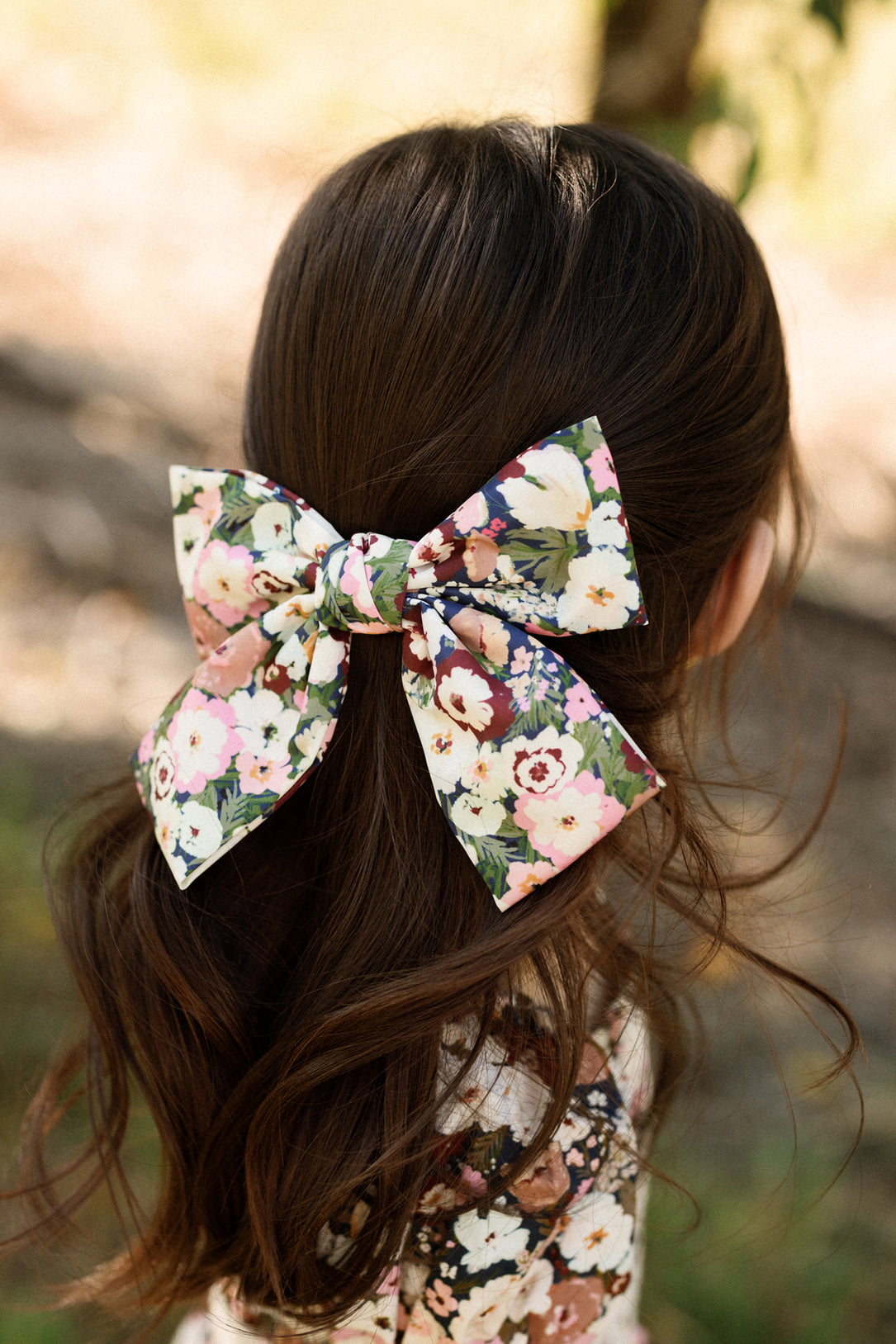 A child with long brown hair wears an Ivy City Co “Hair Bow in Midnight Garden” on the back of their head. The softly blurred background showcases natural outdoor scenery evocative of Ivy City’s enchanting Midnight Garden.