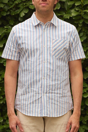 A man stands before green foliage in the Ivy City Co Men's James Shirt in Heritage Stripe—featuring vertical blue, red, and white stripes with a chest pocket—paired with light-colored shorts for a fresh take on classic menswear.