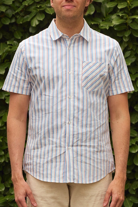 A man stands before green foliage in the Ivy City Co Men's James Shirt in Heritage Stripe—featuring vertical blue, red, and white stripes with a chest pocket—paired with light-colored shorts for a fresh take on classic menswear.
