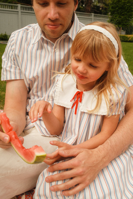 A man wears the Ivy City Co Men's James Shirt in Heritage Stripe while a young girl in a matching dress reaches for his watermelon slice, capturing relaxed, stylish family moments.