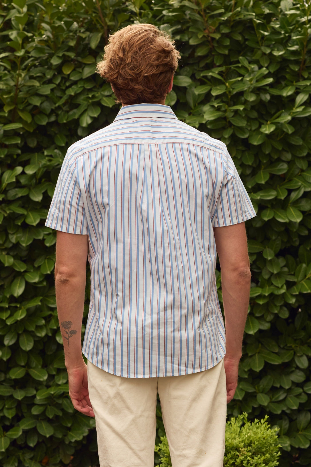 A person with short, wavy hair faces away from the camera in front of leafy green bushes, wearing the Ivy City Co Men's James Shirt in Heritage Stripe and light-colored pants. A small tattoo appears on their left forearm.