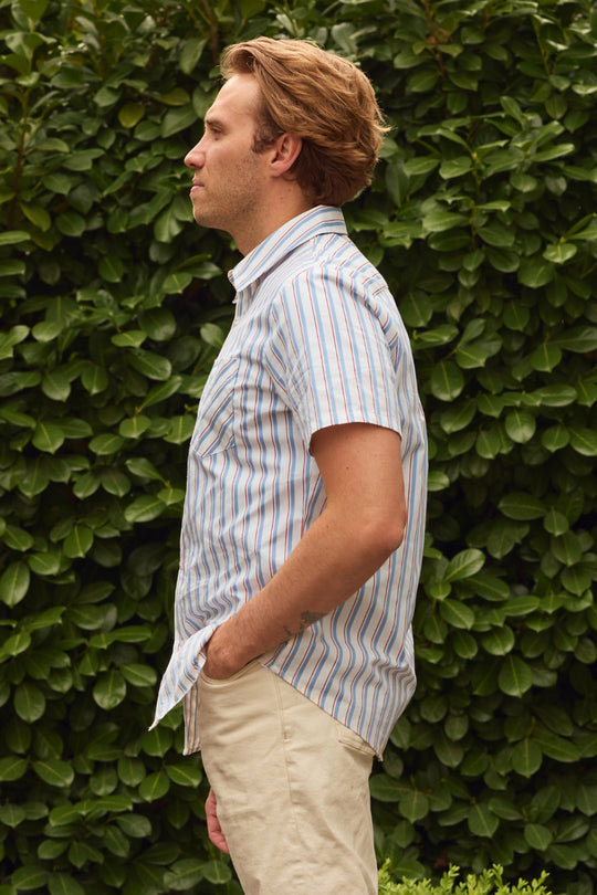 A man in a short-sleeve Ivy City Co Men's James Shirt in Heritage Stripe and light pants stands in profile with hands in pockets before lush greenery—ideal inspiration for effortless men’s style or coordinated family looks.