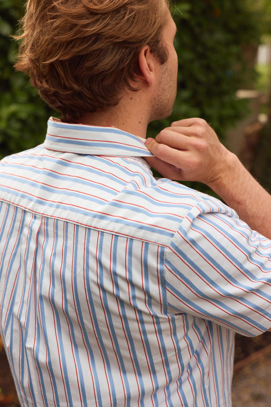 A person with light brown hair, seen from behind, adjusts the collar of the Ivy City Co Men's James Shirt in Heritage Stripe—a light blue shirt with red and white stripes—while standing outdoors near greenery.