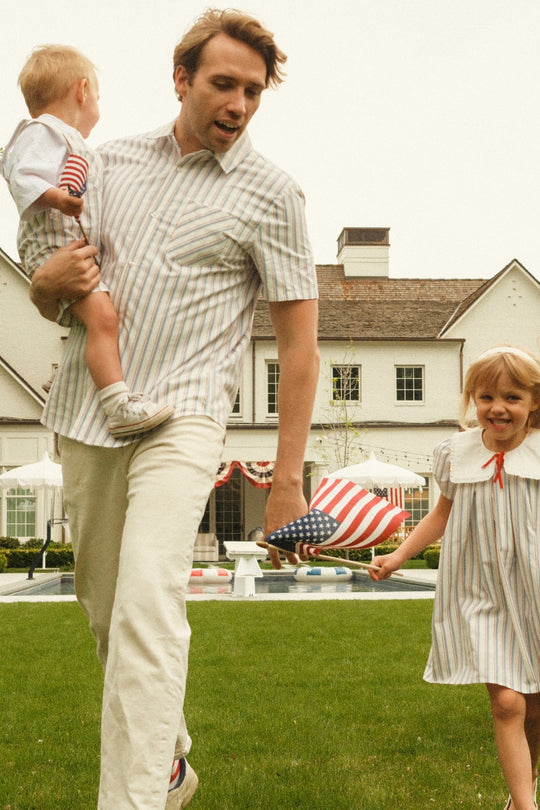 Wearing the Ivy City Co Men's James Shirt in Heritage Stripe, a man holds a child and walks alongside a smiling girl on the lawn of a flag-decorated house, capturing matching family style at a festive summer gathering.
