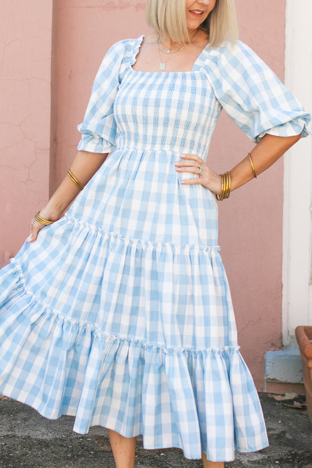 A woman stands in front of a pink wall wearing the Ivy City Co Mia Gingham Dress in Blue, featuring puffed sleeves and a ruffled skirt. This stylish, nursing-friendly dress is perfect for new moms, accessorized with gold bracelets and a necklace.