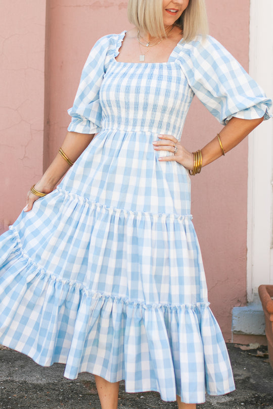 A woman stands in front of a pink wall wearing the Ivy City Co Mia Gingham Dress in Blue, featuring puffed sleeves and a ruffled skirt. This stylish, nursing-friendly dress is perfect for new moms, accessorized with gold bracelets and a necklace.