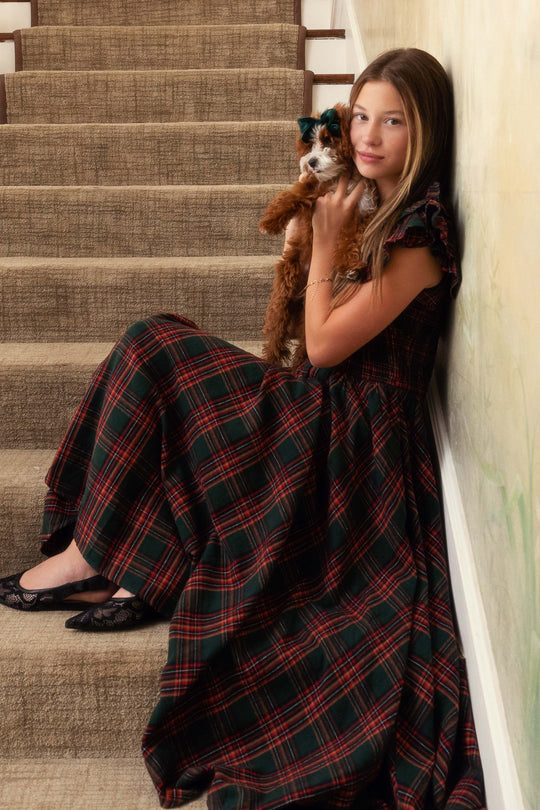 Woman in a plaid dress holding a dog, leaning against a wall on a staircase.