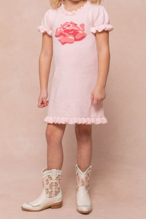 A young child wears the Ivy City Co Mini Rosie Knit Dress, a light pink dress with a rose chest pattern, ruffled sleeves and hem, paired with cream cowboy boots with floral embroidery. The childs face is not shown.