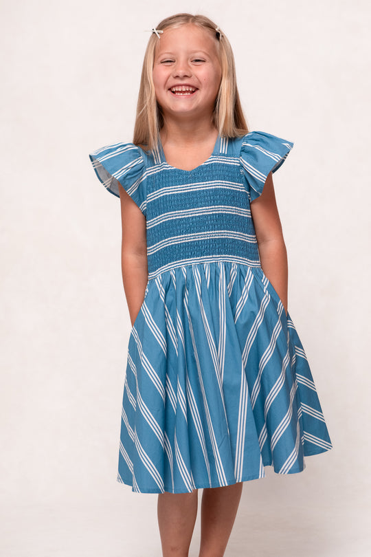 A young girl smiles with hands in pockets, wearing the Ivy City Co Mini Adelaide Dress in Blue—a blue and white striped cotton midi dress with ruffled sleeves—against a plain light background.