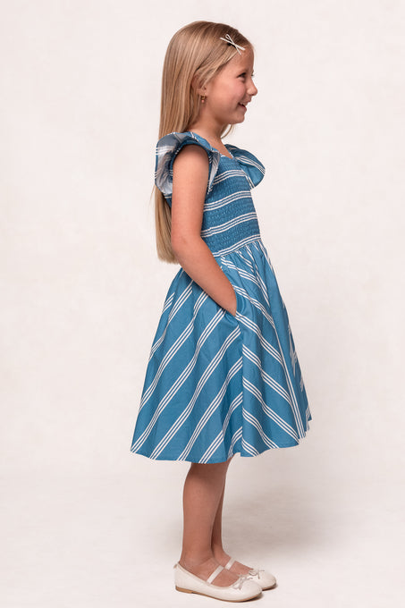 A young girl with long blonde hair smiles in profile, wearing the Ivy City Co Mini Adelaide Dress in Blue—a blue and white striped cotton midi with ruffled sleeves and pockets—paired with white shoes against a plain light background.