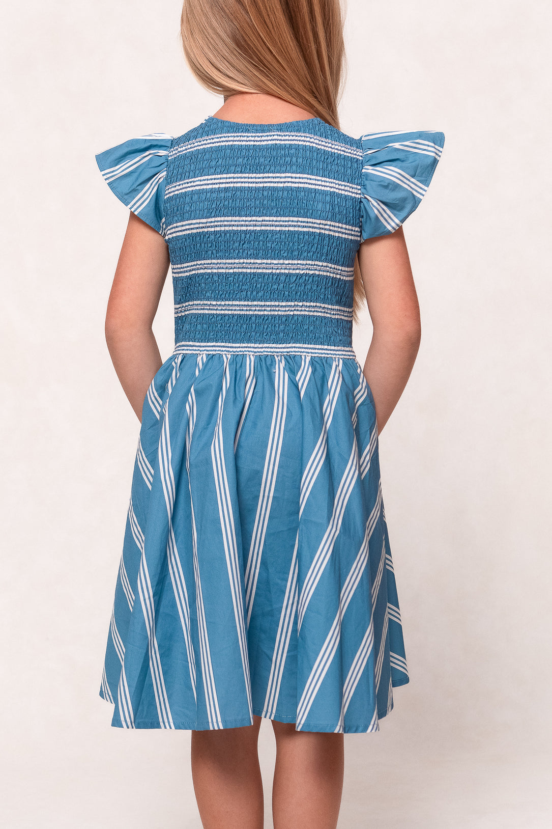 A young girl with long blond hair, shown from behind, wears the Ivy City Co Mini Adelaide Dress in Blue—a cotton midi dress with blue and white stripes, short ruffled sleeves, a smocked bodice, and a skirt that falls just above her knees.