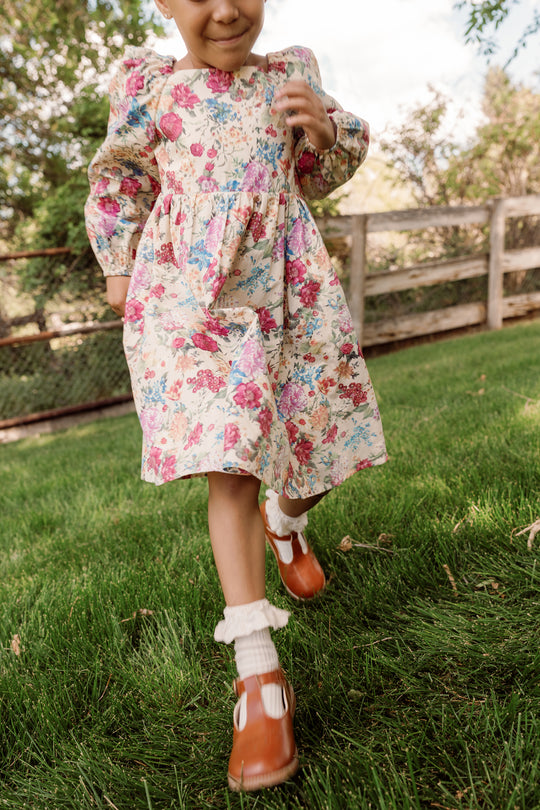 A child wearing the Ivy City Co Mini Adelpha Dress in Autumn Amity Floral, with its smocked bodice and playful balloon sleeves, runs across green grass in a yard framed by a wooden fence and leafy trees.
