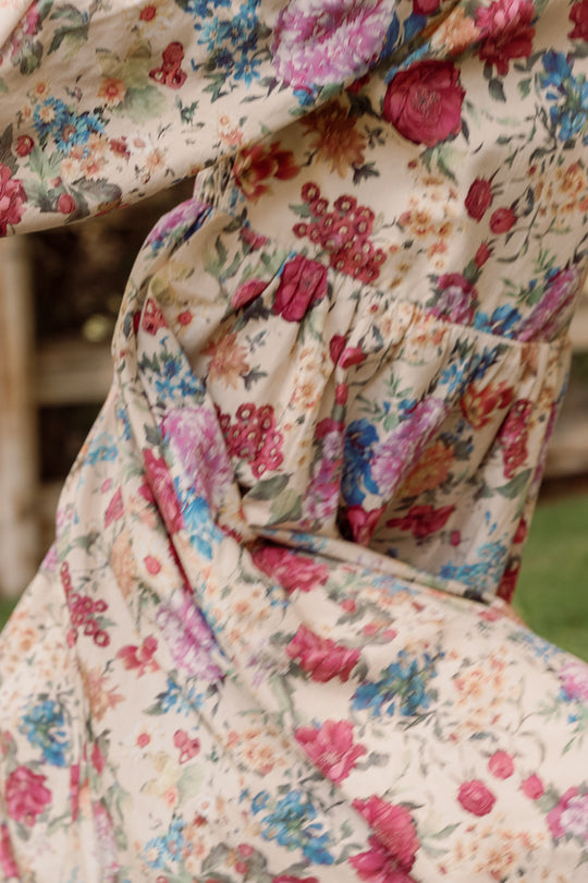 Close-up of the Ivy City Co Mini Adelpha Dress in Autumn Amity Floral, highlighting its flowing fabric as it moves outdoors. The dress boasts a lively floral pattern in shades of pink, red, blue, and green against a beige backdrop. Textured details such as a smocked bodice and delicate balloon sleeves enhance the vibrant appeal of this style.