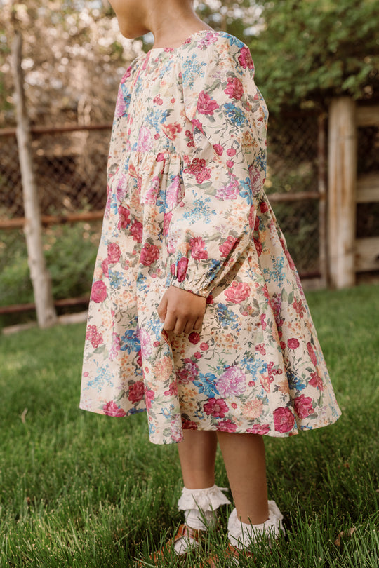 A young girl in the Ivy City Co Mini Adelpha Dress in Autumn Amity Floral, featuring a smocked bodice and balloon sleeves, stands on green grass near a wooden fence and trees with her face turned away from the camera.
