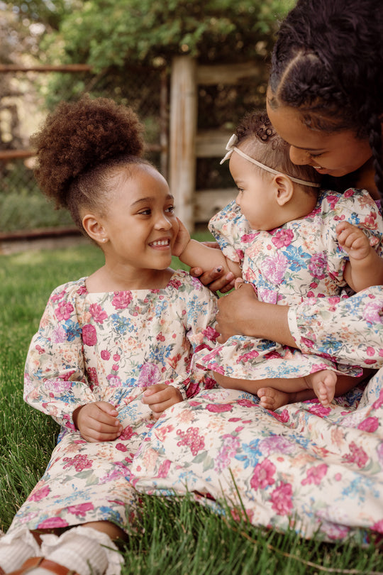 A woman holds a baby and sits beside a young girl, all three smiling at each other while sitting on green grass outdoors. They are wearing matching Ivy City Co Mini Adelpha Dresses in Autumn Amity Floral, featuring playful balloon sleeves and smocked bodice details.