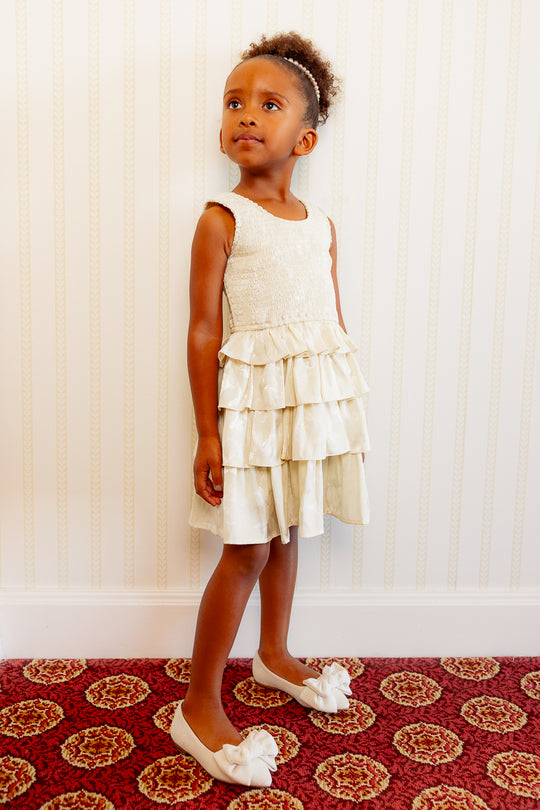 little girl in a white ruffle dress against a white wall and standing on a red carpet.