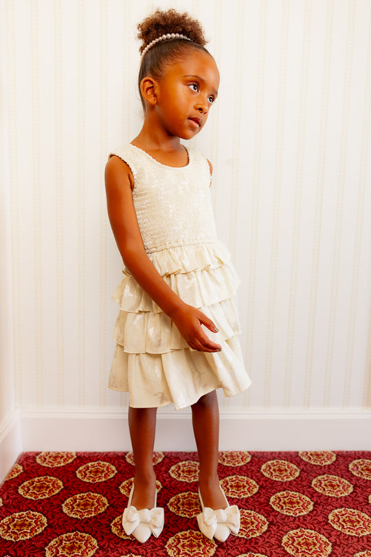 little girl in a white ruffle dress standing against a white wall and standing on a red carpet.