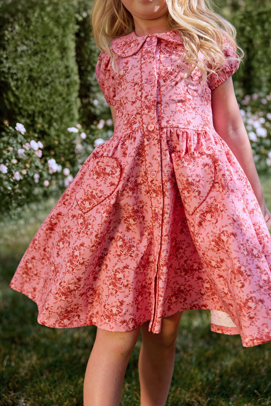 A young girl stands in a garden wearing the Ivy City Co Mini Alayna Play Dress in Desert Heritage Rose. Her blonde hair is loose, and green bushes with white flowers appear in the background.