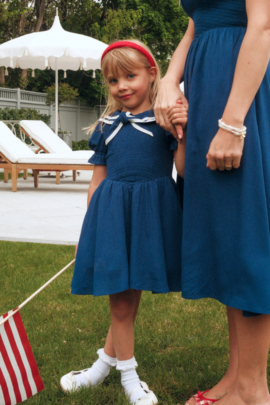 A young girl in an Ivy City Co Mini Alice Dress in Navy and a red headband holds hands with a woman in a matching blue dress. They stand on the grass by poolside lounge chairs, the girl waving an American flag, both wearing sandals.