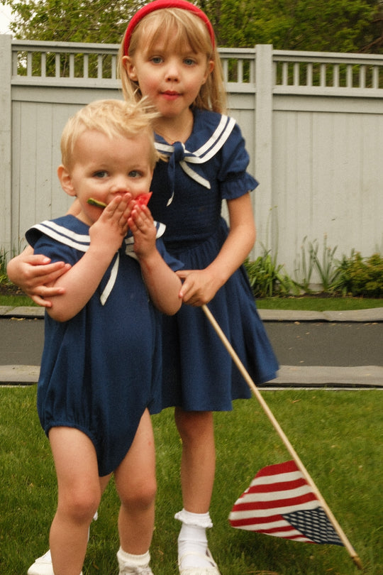 Two young children stand on grass by a fence in matching navy outfits with white sailor collars. The older wears the Ivy City Co Mini Alice Dress in Navy and holds an American flag, while the younger covers their mouth with their hands.