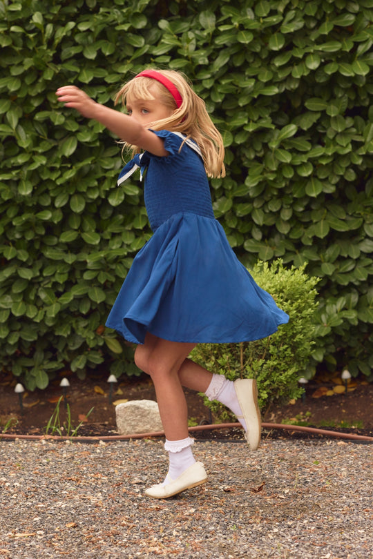 A young girl wearing the Ivy City Co Mini Alice Dress in Navy and a red headband twirls outside on a gravel path, with green bushes behind her.