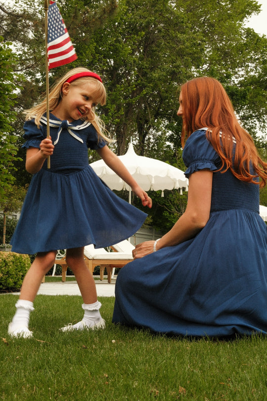 A young girl in the Mini Alice Dress in Navy by Ivy City Co twirls with an American flag, smiling at a woman kneeling beside her; both wear matching blue Ivy City Co outfits on green grass with trees and white umbrellas behind them.
