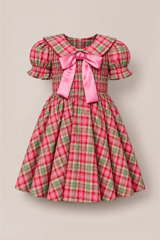 The Ivy City Co Mini Alice Dress in Rosewood Tartan showcases a delightful red, pink, and green plaid pattern with puff sleeves, a smocked bodice, wide collar, and an adorable pink satin bow at the neckline. Its flared skirt adds to the sweet design, making it perfect for mommy-and-me moments. Displayed against a neutral background.