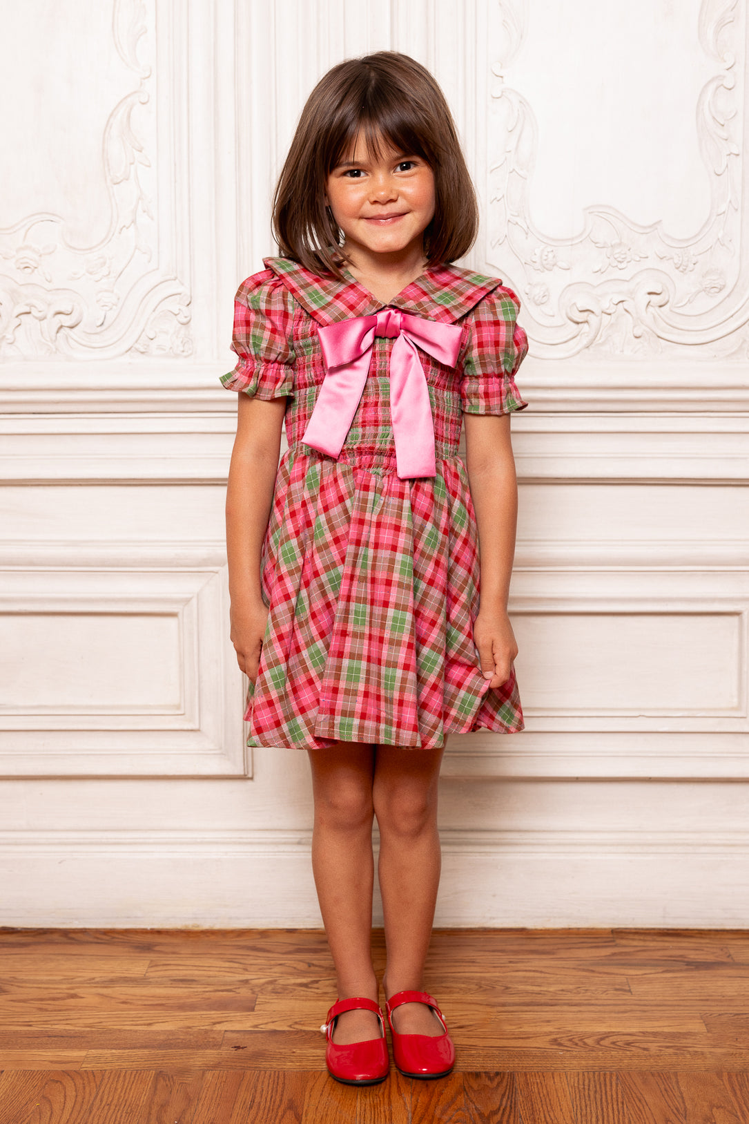 Young girl wearing a red and green plaid dress with a pink bow standing against a white wall.