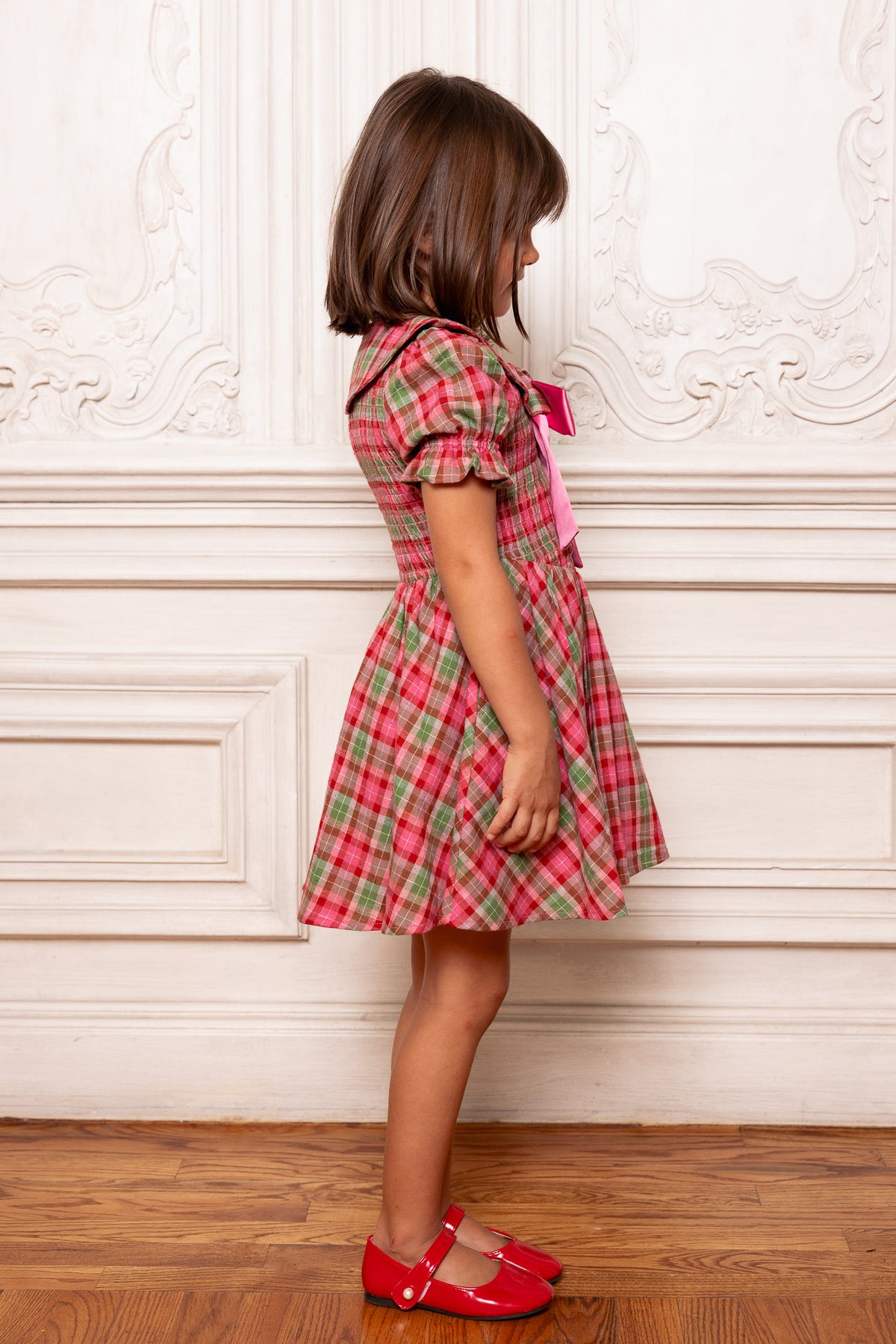Young girl in a red plaid dress standing against a decorative wall.