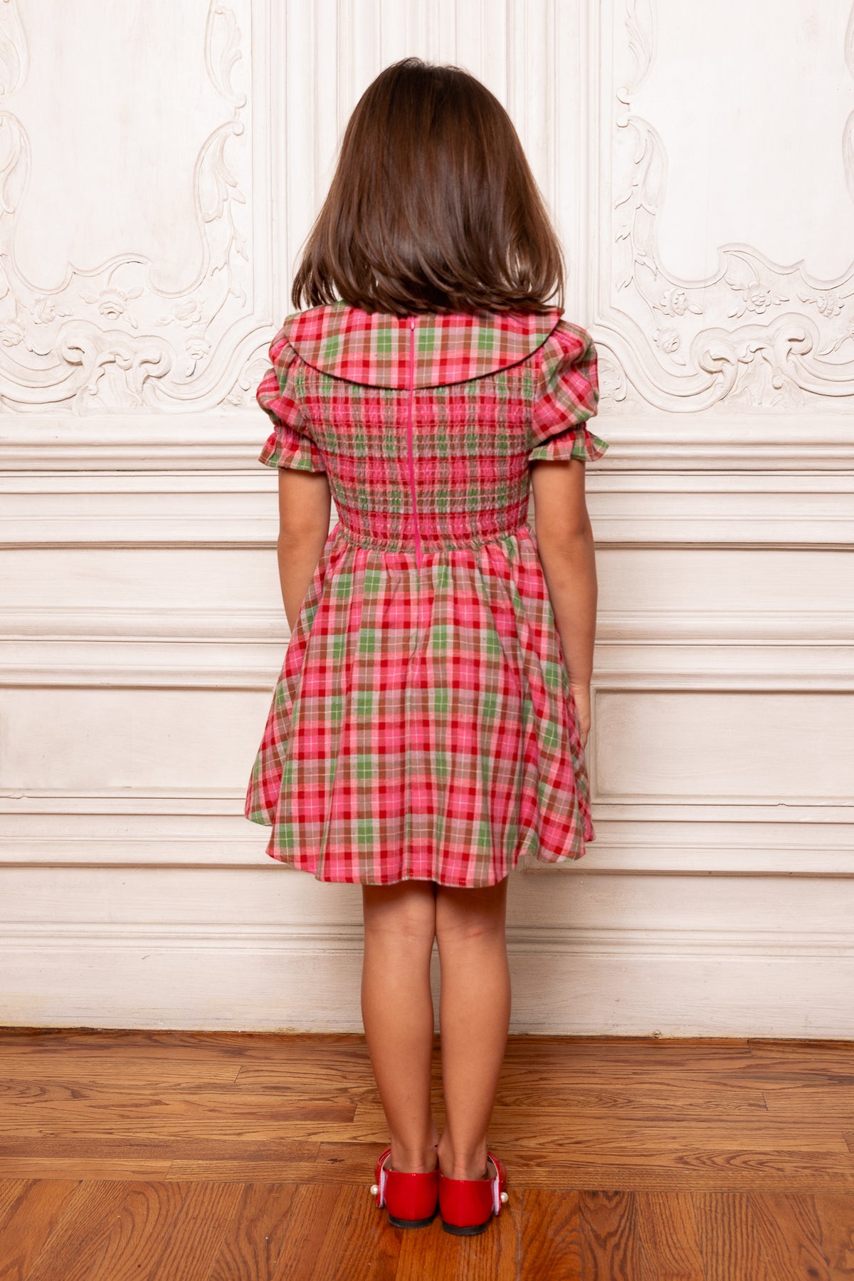 Person wearing a red and green plaid dress standing against a decorative white wall.