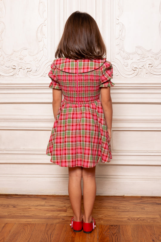 Person wearing a red and green plaid dress standing against a decorative white wall.