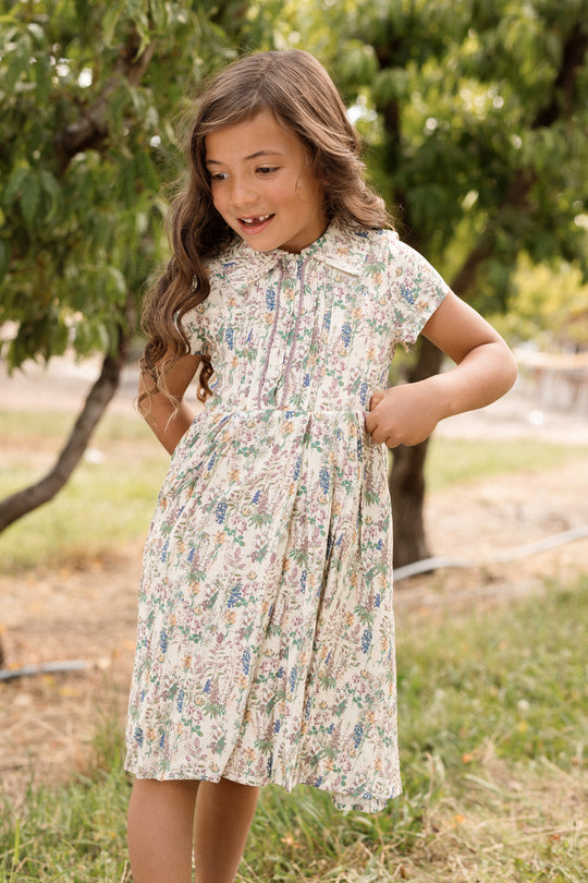 A young girl with long brown hair wears the charming Ivy City Co Mini Amber Dress in Autumn Bouquet, standing outdoors on grass and smiling as she looks to her right, surrounded by trees and lush greenery.
