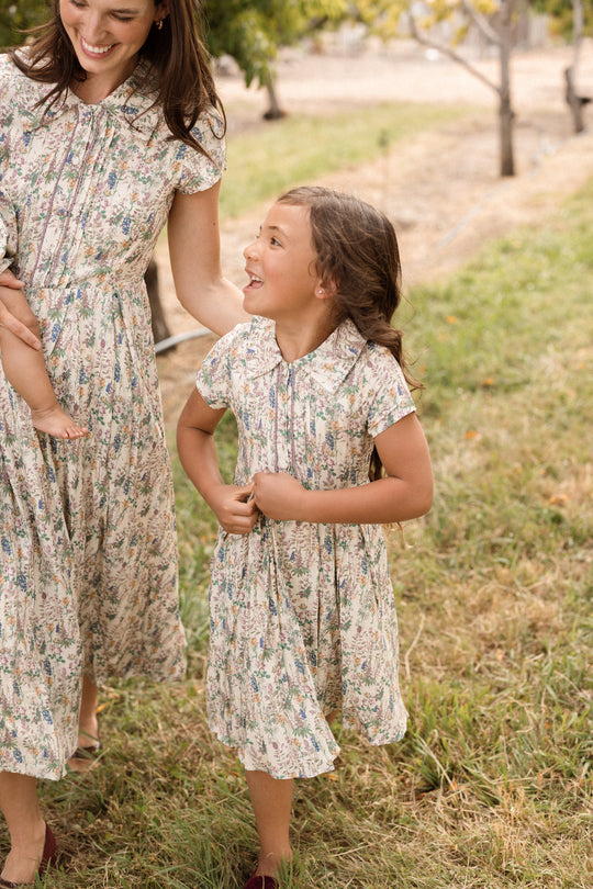 A woman and a young girl, both dressed in matching Ivy City Co Mini Amber Dresses in Autumn Bouquet, smile as they walk together along a grassy outdoor path.