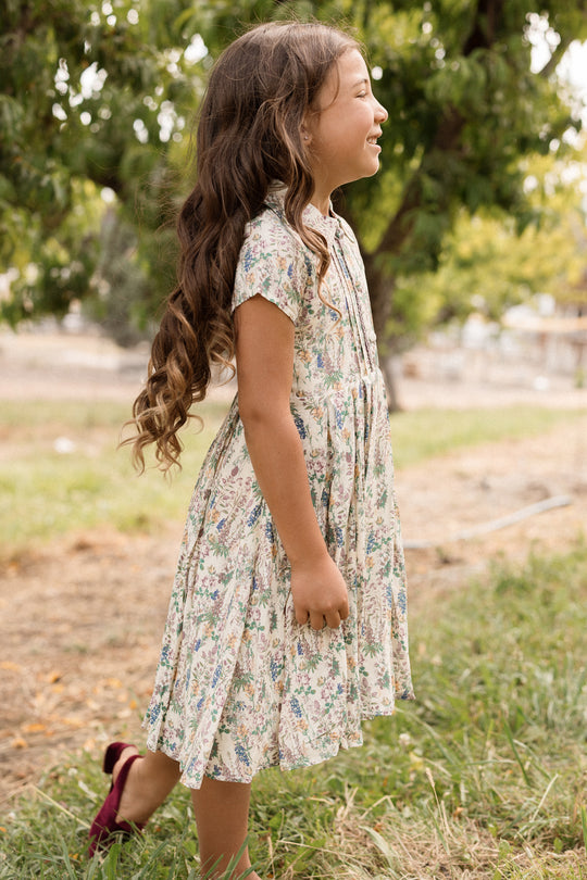 A young girl with long, wavy brown hair, wearing the Ivy City Co Mini Amber Dress in Autumn Bouquet, stands smiling in profile on grass with trees in the background.
