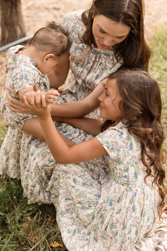 A woman and two young children wearing matching Ivy City Co Mini Amber Dresses in Autumn Bouquet sit on the grass, smiling and playing together outdoors.