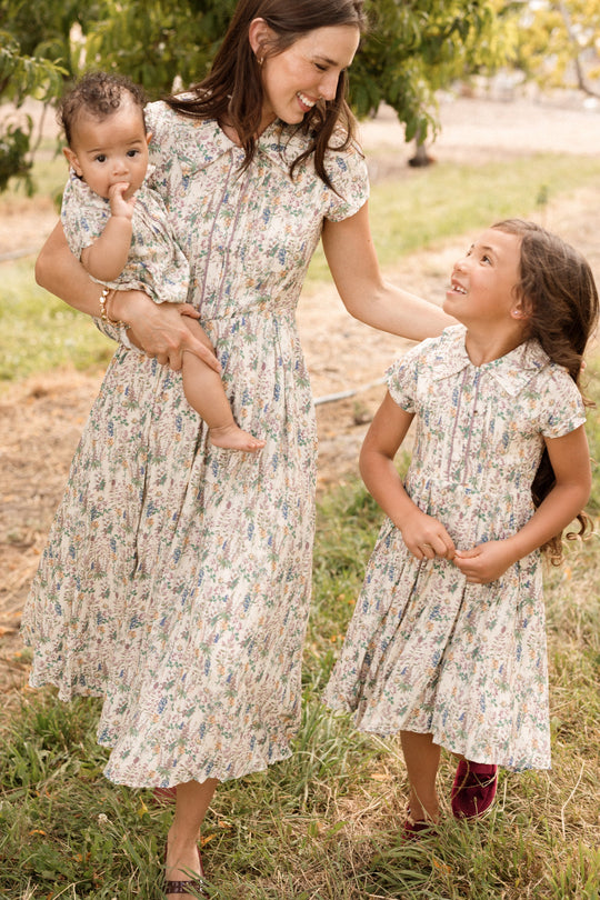 A woman holding a baby and walking with a young girl, all smiling together outdoors on a grassy path, wearing matching Ivy City Co Mini Amber Dresses in Autumn Bouquet.
