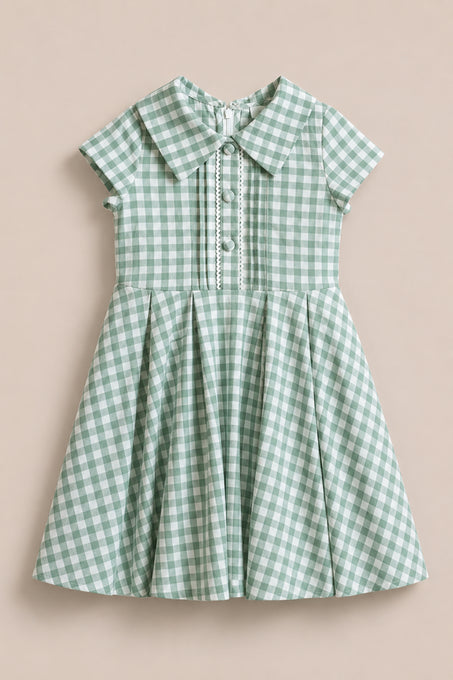The Mini Amber Dress in Sage Gingham by Ivy City Co is a short-sleeved girls' dress featuring a Peter Pan collar, decorative buttons, and a pleated skirt, showcased against a plain beige background.