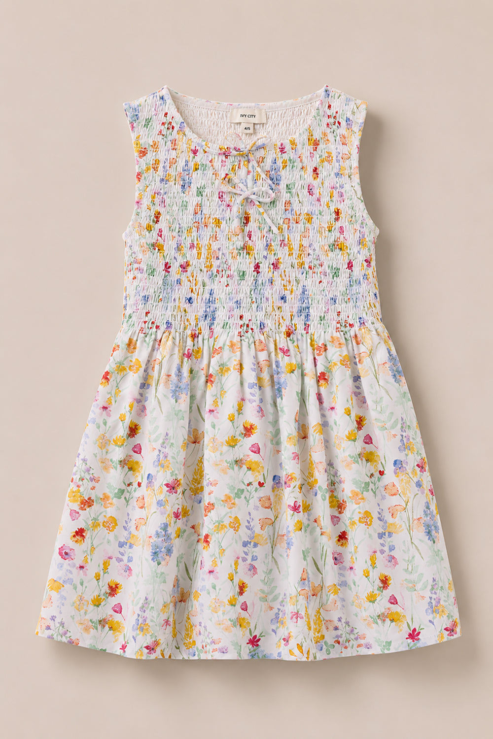 The Mini Ana Dress in Floral Fields by Ivy City Co is a sleeveless, floral print dress for girls with a smocked bodice and a bow at the neckline, shown on a light beige background.