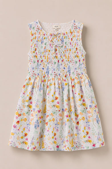 The Mini Ana Dress in Floral Fields by Ivy City Co is a sleeveless, floral print dress for girls with a smocked bodice and a bow at the neckline, shown on a light beige background.