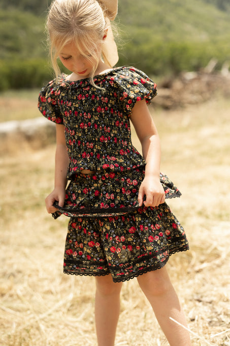 A young blonde girl in a ponytail stands on dry grass, looking down while holding her Ivy City Co Mini Annalise Skirt in Obsidian Vintage Poppies; green hills are visible in the background.