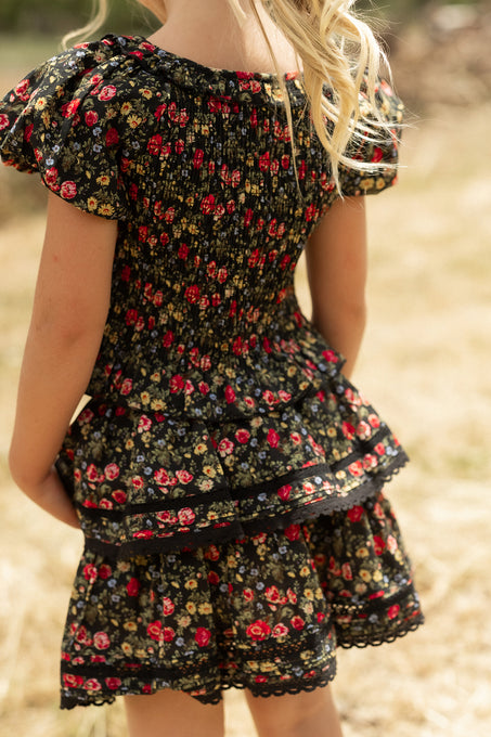 A young blonde girl stands outdoors, shown from the back in a sunny grassy area, wearing the Ivy City Co Mini Annalise Skirt in Obsidian Vintage Poppies with a colorful floral pattern.