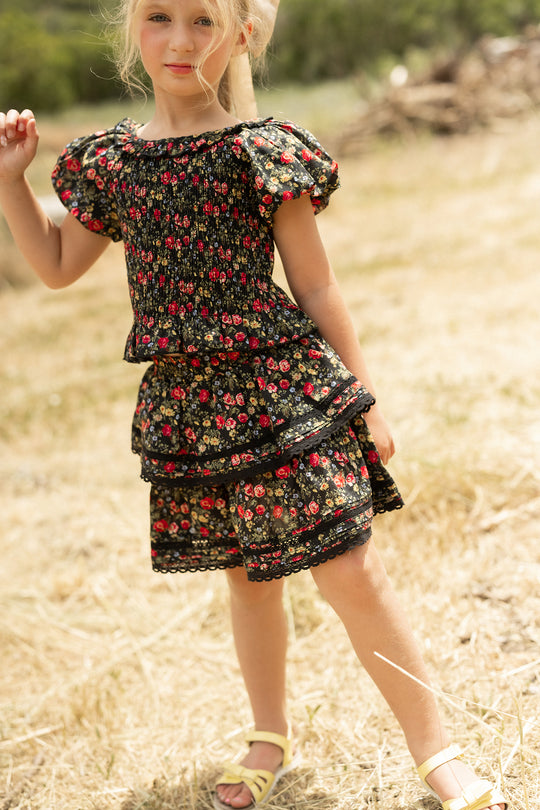 A young girl stands on dry grass, smiling slightly, with her blonde hair tied back. She wears a black floral dress with puffed sleeves, yellow sandals, and the Ivy City Co Mini Annalise Skirt in Obsidian Vintage Poppies.