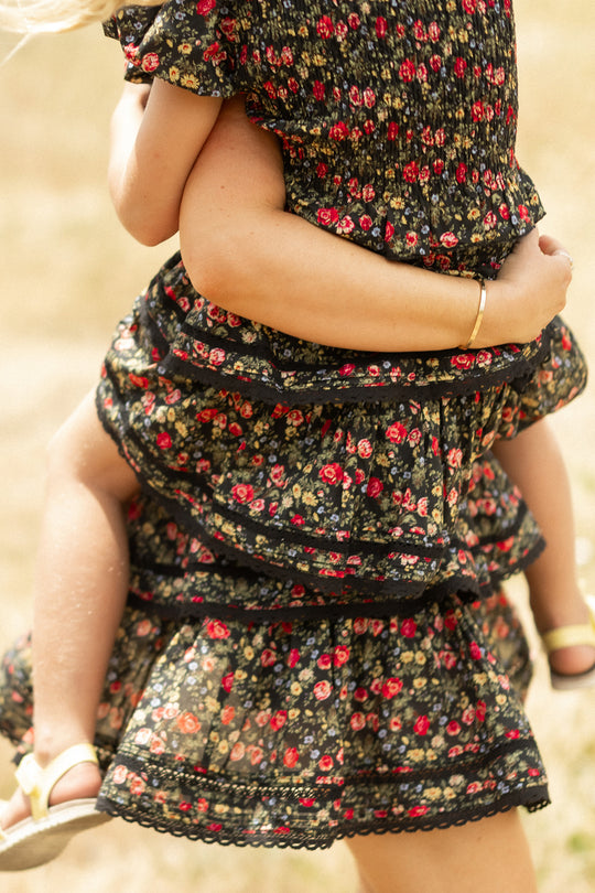 A woman and child enjoy the outdoors in matching black floral dresses; the girl completes her look with yellow sandals and an Ivy City Co Mini Annalise Skirt in Obsidian Vintage Poppies. The scene is bright and summery.