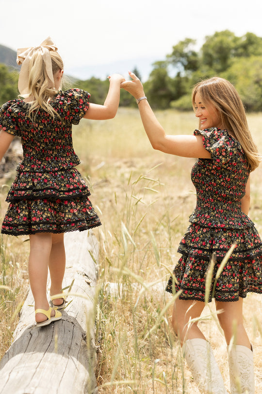 A woman and a girl stroll hand-in-hand in matching Ivy City Co outfits; the girl wears the Mini Annalise Skirt in Obsidian Vintage Poppies with a large hair bow and yellow sandals, while the woman pairs hers with white boots.